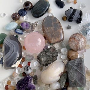 May include: An assortment of polished gemstones and crystals in various shapes and colors. Includes a pink sphere, a carved panda figurine, and a silver pendant with a purple stone. The stones are scattered among small, clear pebbles.
