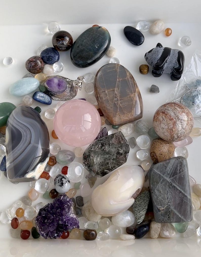 May include: An assortment of polished gemstones and crystals in various shapes and colors. Includes a pink sphere, a carved panda figurine, and a silver pendant with a purple stone. The stones are scattered among small, clear pebbles.