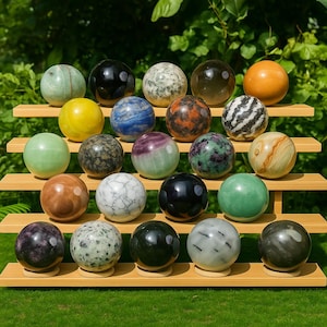 Può includere: Una collezione di ventuno sfere di pietre preziose lucide in vari colori e motivi, esposte su una mensola di bambù a più livelli. Le sfere includono sfumature di verde, nero, marrone, giallo e bianco, con venature e texture uniche.