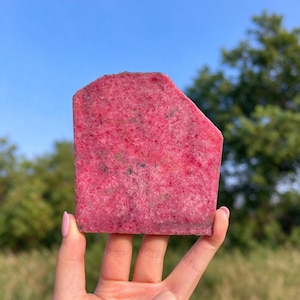 May include: A flat, polished slab of rhodonite mineral featuring a vibrant magenta-pink base color with scattered dark grey and black inclusions throughout the crystalline surface, displaying a natural, irregular geometric shape.