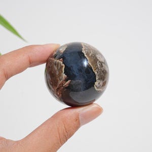 Op de afbeelding: Een gepolijste bolvormige steen met een diepzwarte kern en contrasterende bruine en crèmekleurige minerale insluitingen aan de zijkanten. Het gladde, ronde oppervlak heeft een glanzende afwerking met duidelijke natuurlijke patronen.