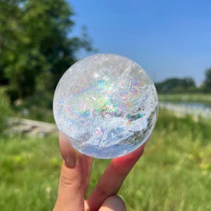Puede incluir: Una bola de cristal esférica y transparente con fracturas internas que crean un vibrante efecto de arcoíris multicolor cuando la luz atraviesa la superficie, mostrando una red compleja de inclusiones prismáticas y reflectantes.