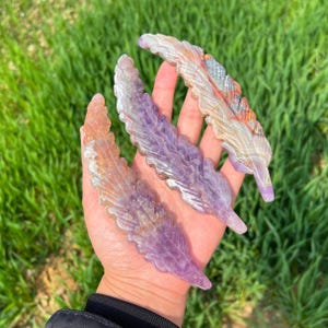 Puede incluir: Tres plumas de piedra tallada en tonos morados, naranjas y blancos se exhiben sobre un fondo de hierba verde. Las plumas tienen vetas detalladas y un acabado pulido, mostrando los colores y patrones naturales de la piedra.