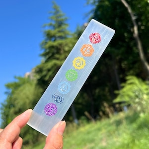 May include: A clear selenite wand, rectangular in shape, featuring seven colourful chakra symbols. The symbols are red, orange, yellow, green, blue, indigo, and purple. The wand is held against a backdrop of green grass and a blue sky.