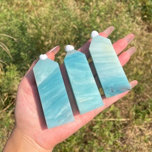 May include: Three polished, light blue amazonite crystal towers with smooth, rectangular bodies and pointed, faceted tops. Each stone features natural white and teal banding, showcasing a translucent, marbled texture throughout the mineral structure.