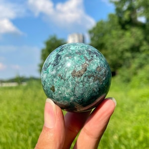 Puede incluir: Una piedra esférica pulida que presenta una textura moteada con inclusiones minerales en verde bosque profundo, azul verdoso y blanco. La superficie muestra un acabado brillante con motas metálicas dispersas que crean un patrón complejo.
