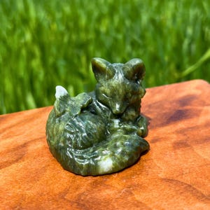 May include: A green fox figurine carved from stone, curled up in a resting pose. The fox has detailed fur and facial features. It sits on a wooden surface, with a blurred green background. The figurine is approximately 7 cm long.