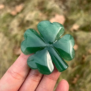 Puede incluir: Un trébol de cuatro hojas pulido, tallado en piedra preciosa verde. El trébol tiene lóbulos redondeados y un acabado brillante. La imagen está tomada al aire libre con un fondo borroso de hierba y follaje.