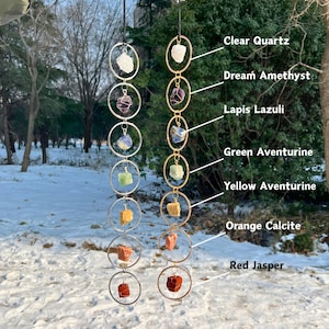 May include: Two hanging crystal suncatchers with various gemstones, including Clear Quartz, Dream Amethyst, and Red Jasper. The crystals are suspended within silver and gold metal rings. Text labels identify each crystal type.