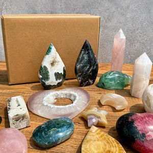 Pode incluir: Uma coleção de pedras preciosas e cristais polidos em várias formas e cores, incluindo gotas, corações e pontas. A variedade apresenta pedras verdes, brancas, rosas e transparentes, juntamente com uma pequena figura de tartaruga verde. Uma caixa de cartão castanha está no fundo.
