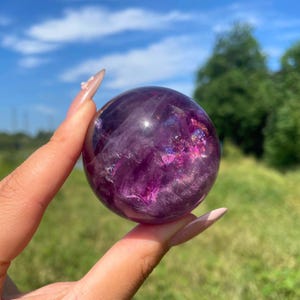 May include: A polished spherical crystal in deep shades of purple and violet. The stone features internal fractures and inclusions that create reflective, iridescent flashes of light throughout its translucent, rounded surface.