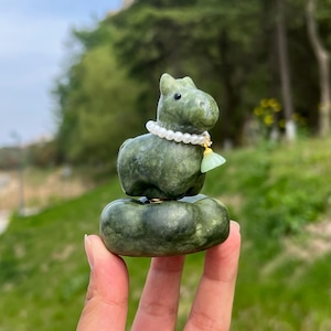 Könnte beinhalten: Eine grüne Jade-Figur eines pferdeähnlichen Tieres mit einer Perlenkette und einer grünen Quaste. Die Figur sitzt auf einem runden, grünen Sockel. Die Skulptur wird von einer Hand gehalten.
