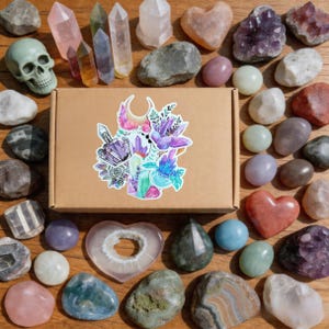May include: An assortment of crystals, gemstones, and a skull arranged on a wooden surface. The collection includes quartz points, heart-shaped stones, and a box with a floral and crystal sticker. Colors include pink, purple, green, and brown.