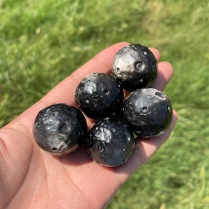May include: Five small, spherical objects with a dark, metallic grey finish and a textured surface featuring numerous circular indentations and craters, resembling the appearance of the moon, held together in a cluster.