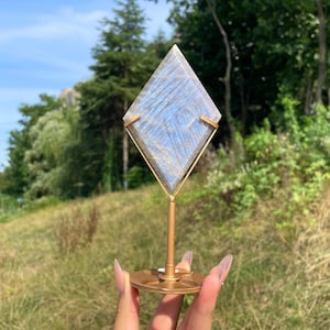 May include: A diamond-shaped labradorite stone with blue and grey iridescent flashes, held in a gold-toned metal stand. The stone features a polished surface with visible natural striations and is mounted on a circular base with a thin stem.