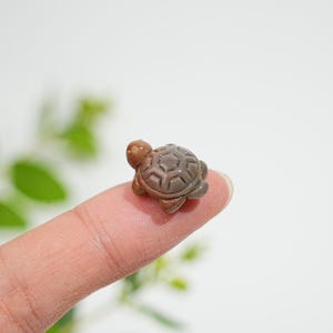 Op de afbeelding: Een klein, uit bruine steen gesneden schildpadfiguurtje met een gladde, gepolijste afwerking en een gedetailleerd zeshoekig schildpatroon. Het miniatuurbeeldje heeft een afgeronde kop en vier kleine pootjes met natuurlijke kleurvariaties.