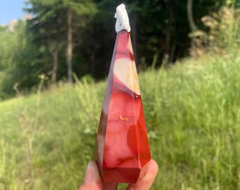 May include: A red, white, and beige gemstone obelisk with a carved white figure at the top. The obelisk is held in a hand against a backdrop of green grass and trees. The obelisk is approximately 6 inches tall.