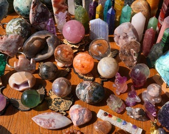 May include: A collection of various polished crystal spheres, towers, and carved animal figurines in shades of pink, green, blue, and brown, alongside several beaded bracelets and a small rectangular white stone bar with seven colored inlaid stones.
