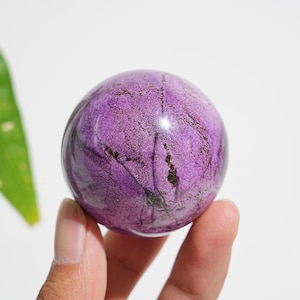 May include: A polished spherical stone with a vibrant purple hue, featuring intricate dark brown and black veining throughout its surface. The sphere displays a smooth, glossy texture with natural mineral inclusions that create a unique pattern.