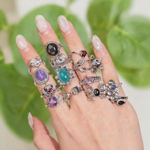 May include: A hand adorned with numerous silver rings, each featuring different gemstones. The rings showcase black, purple, teal, and clear stones, alongside decorative elements such as hearts, bows, and animal motifs. The backdrop is a gentle green.