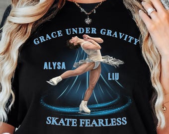 May include: A black t-shirt featuring a graphic of a figure skater in a white dress. The text reads GRACE UNDER GRAVITY, ALYSA LIU, and SKATE FEARLESS in light blue lettering. The design includes blue light streaks and a circular motion effect.