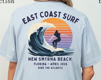 May include: A light blue t-shirt featuring a graphic on the back with the text EAST COAST SURF, NEW SMYRNA BEACH, FLORIDA, APRIL 2026, RIDE THE ATLANTIC. The design shows a surfer on a wave against a sunset of orange and purple.