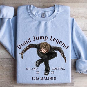 May include: A light blue sweatshirt featuring a graphic of a figure skater in a dynamic pose. The text "Quad Jump Legend" arches above the skater, with "MILANO 20" and "CORTINA 26" below. The name "ILIA MALININ" is printed at the bottom.