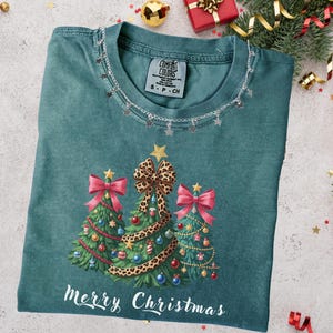 May include: Teal t-shirt featuring a Christmas tree design. The design includes three decorated Christmas trees with bows, ornaments, and a leopard print bow. The text "Merry Christmas" is printed at the bottom. A silver chain with star charms decorates the neckline.