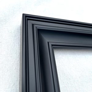 May include: A matte black picture frame featuring a multi-tiered, stepped profile design. The frame has a clean, angular corner joint and a smooth, uniform finish across its entire surface, highlighting the depth and structure of the molding.