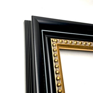 May include: A rectangular picture frame featuring a wide, smooth matte black outer border with a stepped profile. The inner edge is accented by a gold-toned trim decorated with a raised, repeating bead pattern that adds texture to the frame.
