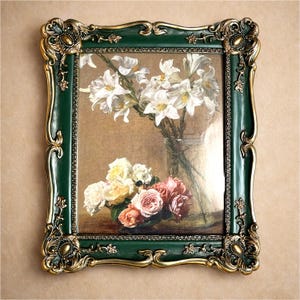 May include: A framed floral still life. The painting depicts white lilies and a bouquet of roses in a glass vase. The frame is dark green with gold accents and ornate detailing, giving a vintage aesthetic.