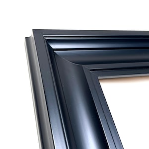 May include: A black picture frame with a smooth, matte finish and a multi-tiered, stepped profile. The frame features a deep, concave inner edge that transitions into a series of sharp, linear ridges, creating a structured, layered appearance.
