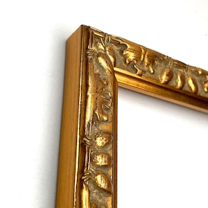 May include: A gold-coloured picture frame featuring a raised relief pattern of acorns and oak leaves along the inner border. The frame has a smooth outer edge and a textured, metallic finish, showcasing a corner section of the rectangular structure.