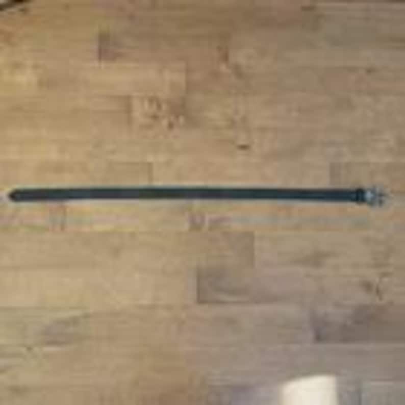 Op de afbeelding: Een zwarte leren riem met een zilverkleurige gesp ligt op een lichtbruin houten oppervlak. De riem is lang en dun, met een klassiek ontwerp. De gesp is rechthoekig en heeft een enkele doorn.