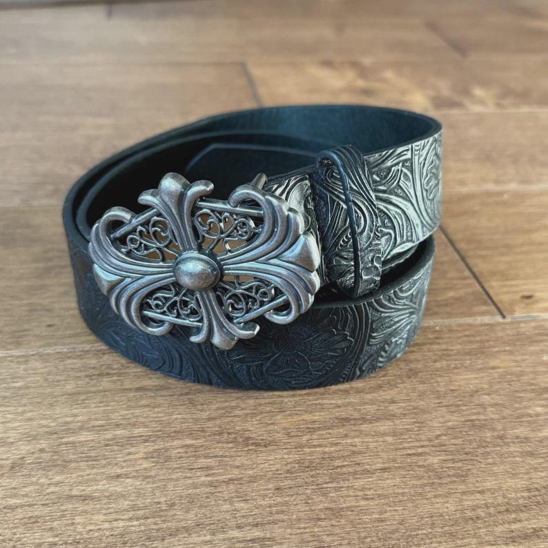 Op de afbeelding: Zwarte leren riem met een zilverkleurige, sierlijke gesp. De riem heeft een bloemreli&euml;fpatroon. De gesp is een gedetailleerd, decoratief ontwerp. De riem ligt op een houten oppervlak.