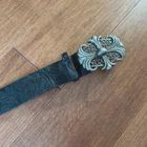 Op de afbeelding: Een zwarte leren riem met een zilverkleurige, sierlijke gesp. De gesp heeft een gedetailleerd, symmetrisch ontwerp met een kruisachtig element in het midden. De riem lijkt versleten, met een getextureerd oppervlak.