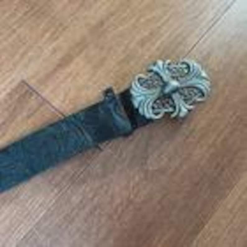 Op de afbeelding: Een zwarte leren riem met een zilverkleurige, sierlijke gesp. De gesp heeft een gedetailleerd, symmetrisch ontwerp met een kruisachtig element in het midden. De riem lijkt versleten, met een getextureerd oppervlak.