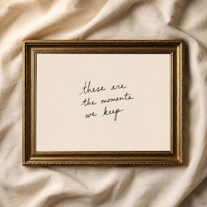May include: A rectangular wall art piece featuring a metallic gold frame with a textured border. Inside, a cream-colored background displays the black handwritten text: these are the moments we keep. The design is centered within the frame.