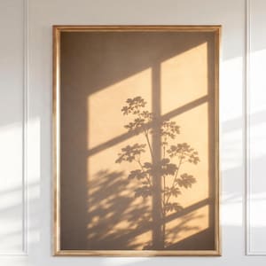 May include: A rectangular wall art piece featuring a gold frame surrounding a print of a warm, tan-colored wall surface. The image displays the dark, crisp shadow of a leafy plant and the geometric shadow of a window frame cast across the surface.