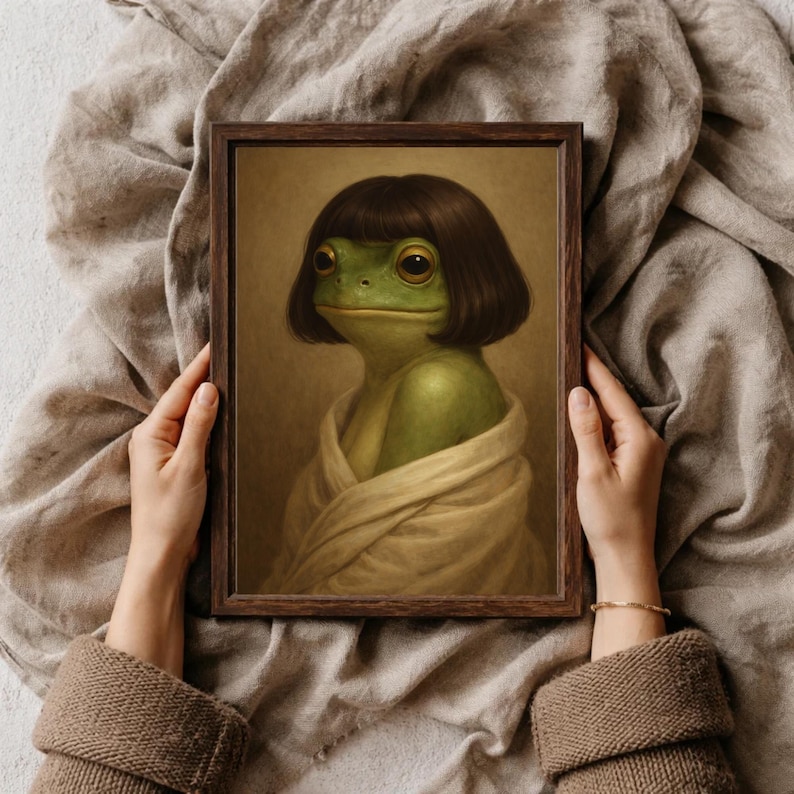 以下が含まれることがあります： ボブヘアの緑色のカエルとクリーム色のラップを描いた額入りのアートプリント。カエルは大きく表現力豊かな目を持ち、古典的な肖像画スタイルで描かれています。フレームは濃い茶色です。