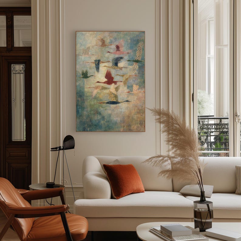 May include: An abstract painting featuring a flock of birds in flight. The birds are depicted with various colors, including red, blue, and yellow, against a textured, muted background. The artwork is framed and displayed above a white sofa.