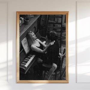 Könnte beinhalten: Schwarz-Weiß-Druck in einem Rahmen, der ein Paar an einem Klavier zeigt. Die Frau lehnt sich an das Klavier, während der Mann daneben sitzt. Klaviertasten und Noten sind sichtbar. Der Druck befindet sich in einem goldfarbenen Rahmen.
