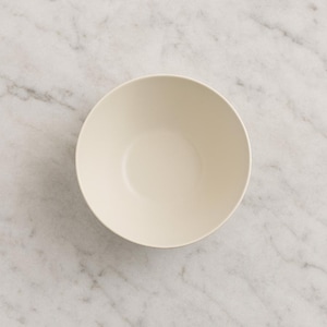 May include: A cream-colored ceramic bowl with a simple, rounded design. The bowl has a smooth, matte finish and a wide, shallow shape. The bowl is empty and sits on a marble surface.