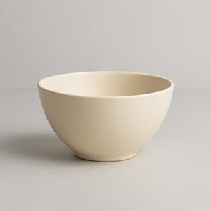 May include: A cream-colored ceramic bowl with a rounded shape. The bowl has a smooth, matte finish and a simple, minimalist design. The bowl is suitable for serving soups, cereals, or other dishes.