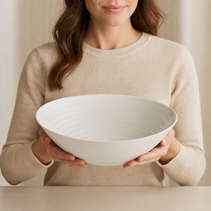 May include: A large, white ceramic bowl with a textured, ridged design. The bowl is held by a person wearing a beige sweater. The bowl's shape is wide and shallow, suitable for serving salads or displaying fruit.