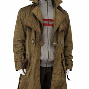 Può includere: Un lungo trench coat verde oliva con ampio colletto e dettagli con bottoni. Indossato sopra una giacca grigia con zip e due strisce rosse sul petto. Il manichino indossa anche pantaloni neri.