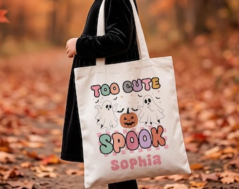 May include: A white canvas tote bag featuring a colorful graphic with two ghosts, a jack-o'-lantern, and bats. The text reads "TOO CUTE SPOOK" in retro-style lettering with the name "Sophia" printed in pink below the main design.