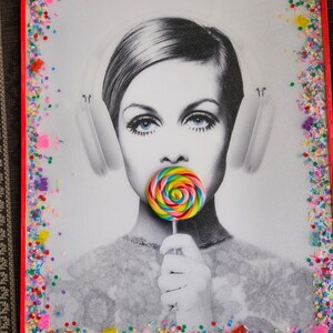 May include: A black and white photo of a woman wearing headphones and holding a colorful lollipop. The photo is framed with a red frame and a border of colorful sprinkles.