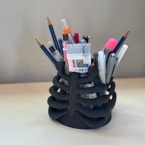 May include: A matte black pencil holder shaped like a human rib cage, filled with various writing instruments including pencils, pens, and markers. One pen features a label with the Japanese text ノック式 and 0.5, indicating a retractable mechanism.