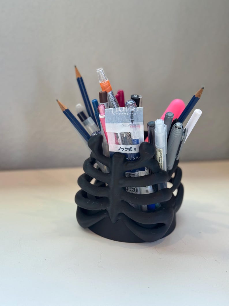 May include: A matte black pencil holder shaped like a human rib cage, filled with various writing instruments including pencils, pens, and markers. One pen features a label with the Japanese text ノック式 and 0.5, indicating a retractable mechanism.
